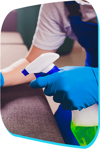 A person wearing blue rubber gloves is holding a spray bottle with a green liquid, spraying it onto a fabric surface, likely for cleaning purposes.