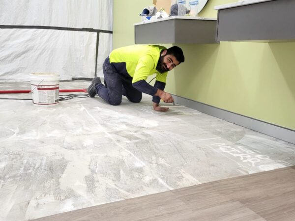 A man in a neon yellow shirt kneels on the floor while smoothing a layer of material, likely working on a flooring project.