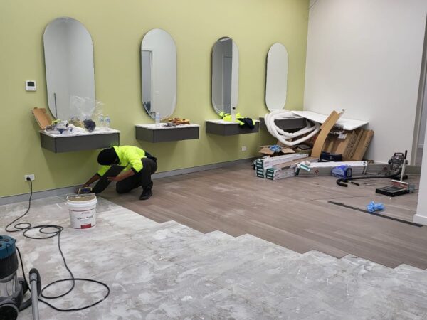 A partially renovated room with a lime green accent wall featuring three oval mirrors mounted above floating countertops. A worker in a high-visibility shirt is kneeling on the floor, working on laying wood flooring. The room has recessed ceiling lights and exposed ventilation ducts. The floor is partially covered with unfinished concrete and partially with newly installed wooden planks. Construction materials and tools are scattered throughout the space, including a vacuum cleaner, a bucket, and various packaging materials.