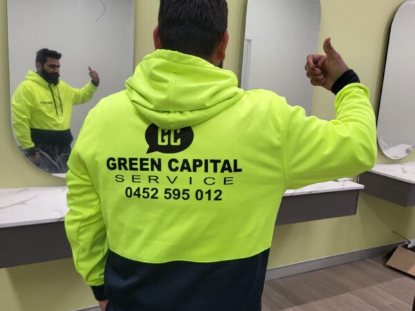 A man wearing a neon yellow hoodie with the company name "Green Capital Service" and a contact number on the back, giving a thumbs-up while standing in front of mirrors.