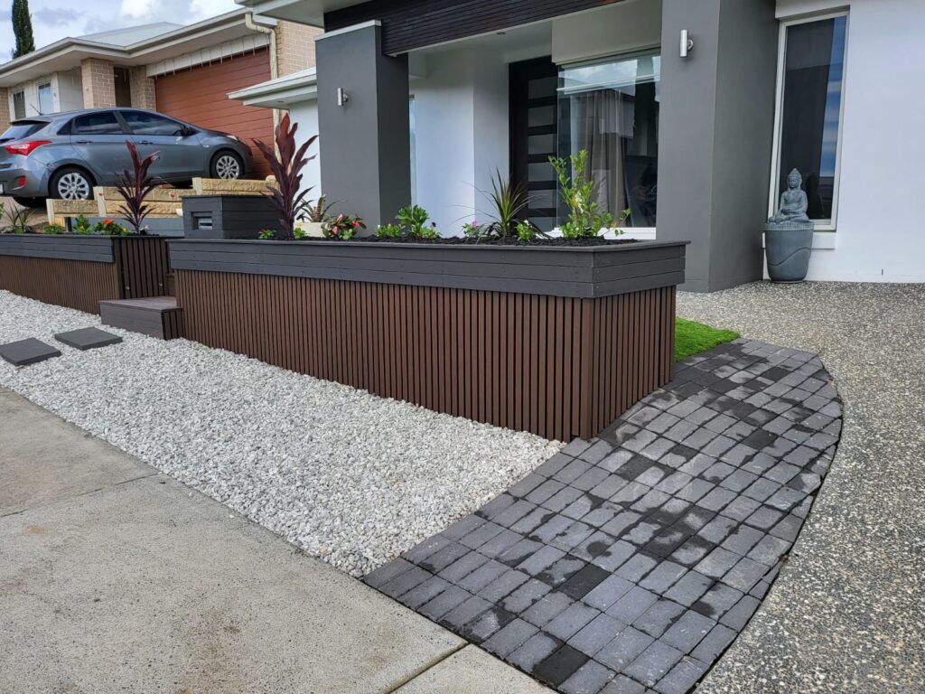 A modern front yard with a raised garden bed featuring horizontal wooden slats and a dark trim. The garden bed is filled with small green plants and bordered by a white gravel section. A pathway of dark gray pavers curves from the driveway to the entrance of the house. The house exterior has a contemporary design with a gray and white color scheme, a large window, and a sleek black front door. A parked car is visible in the background along with neighboring houses.