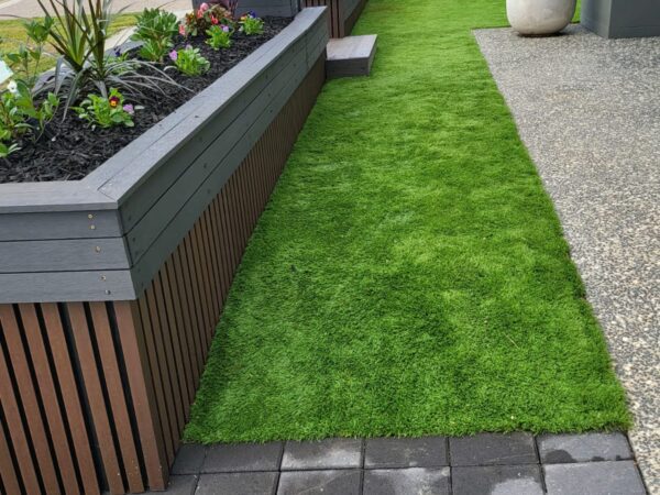 A modern front yard with a raised garden bed featuring horizontal wooden slats and a dark trim. The garden bed is filled with small green plants and bordered by a white gravel section. A pathway of dark gray pavers curves from the driveway to the entrance of the house. The house exterior has a contemporary design with a gray and white color scheme, a large window, and a sleek black front door. A parked car is visible in the background along with neighboring houses